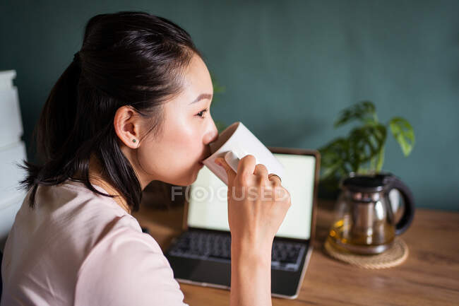 Side view of Asian female freelancer reading documents on laptop while sitting and drinking tea looking away at table during remote work — Stock Photo