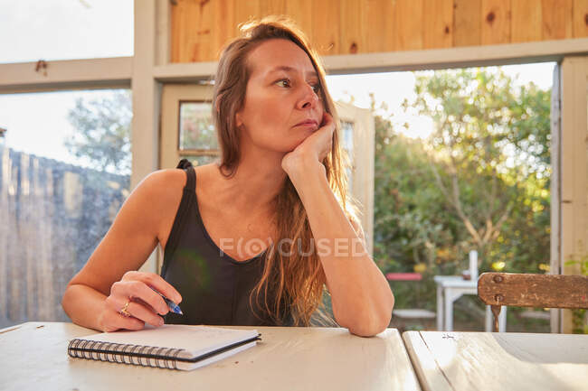 Low angle of cheerful female gardener sitting at table and writing in notepad in greenhouse — Stock Photo