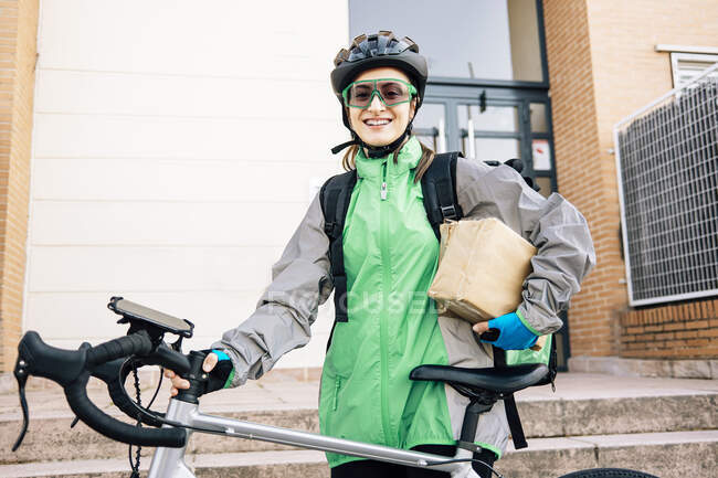 Щаслива жінка, що носить загорнуті коробки і стоїть на вулиці з мотоциклом в сонячний день, дивлячись на камеру — стокове фото