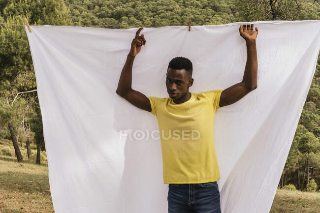Serious African American male model standing on background of white natural textile hanging on clothesline in nature — Stock Photo