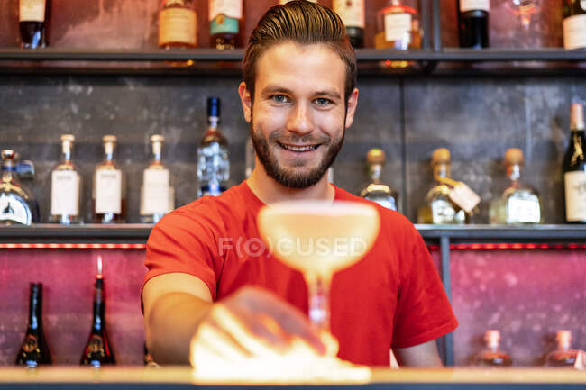 Barman alegre servindo vidro com coquetel de álcool no balcão no bar e olhando para a câmera — Fotografia de Stock
