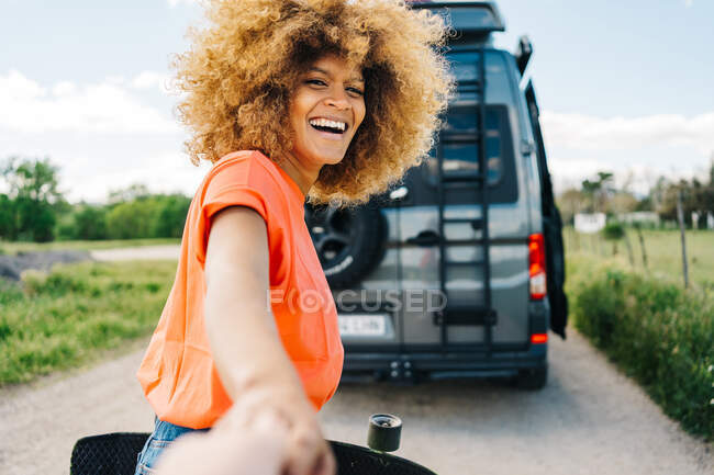 Fröhliche Afroamerikanerin mit Longboard lächelt und blickt in die Kamera, während sie ihren Freund während eines Roadtrips zum Transporter führt — Stockfoto