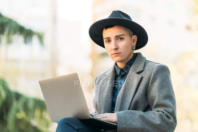 Androgynous persona con mohawk en botas y abrigo navegar por Internet en netbook mientras está sentado en la ciudad - foto de stock