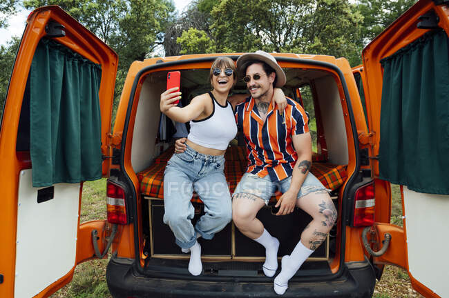 Encantada pareja de viajeros sentados en furgoneta y tomando fotos en el teléfono inteligente mientras abrazan y disfrutan de la aventura de verano - foto de stock