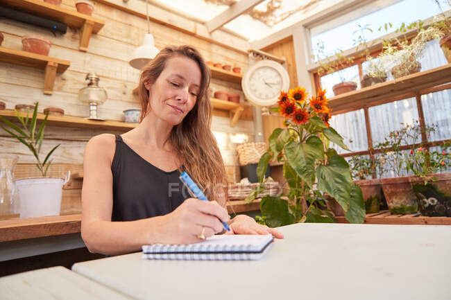 Low angle of cheerful female gardener sitting at table and writing in notepad in greenhouse — Stock Photo