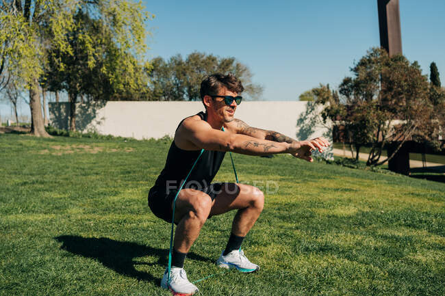 Athlète masculin fort en vêtements de sport et lunettes de soleil s'exerçant avec une bande élastique sur la pelouse à la lumière du soleil — Photo de stock