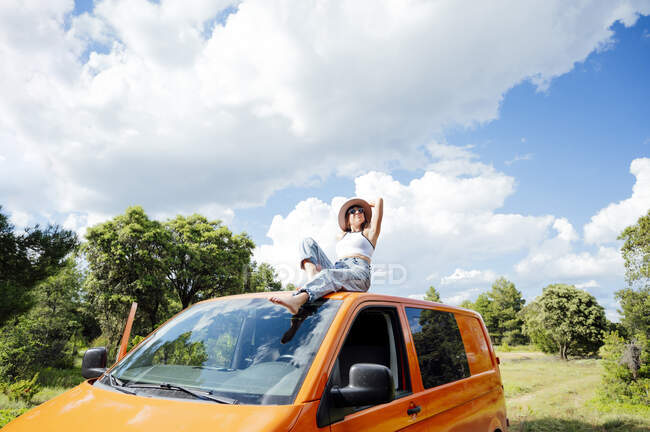 Exploradora femenina serena sentada en el techo de la furgoneta y disfrutando de la aventura de verano en un día soleado - foto de stock