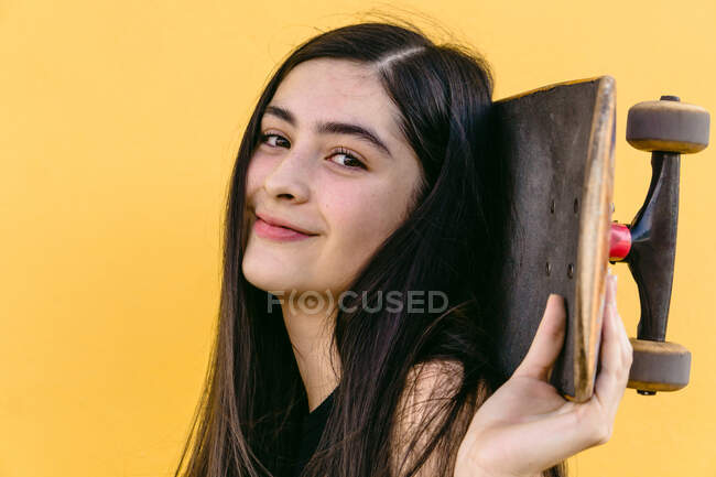 Vista laterale del giovane skateboarder femminile allegro che tiene lo skateboard dietro la testa mentre guarda la fotocamera — Foto stock