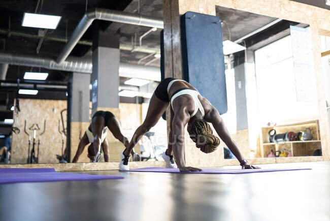 Ground level of anonymous ethnic sportswoman standing in Downward Facing Dog pose during training on mat against mirror in gym — Stock Photo