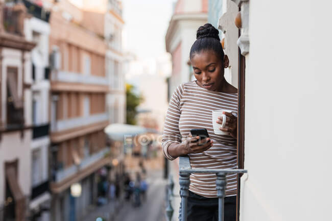 African American female with cup of hot drink surfing internet on cellphone on balcony — Stock Photo