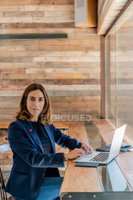 Adult female in elegant outfit surfing on laptop while sitting in chair at table next to notebook in bright cafeteria near window — Stock Photo