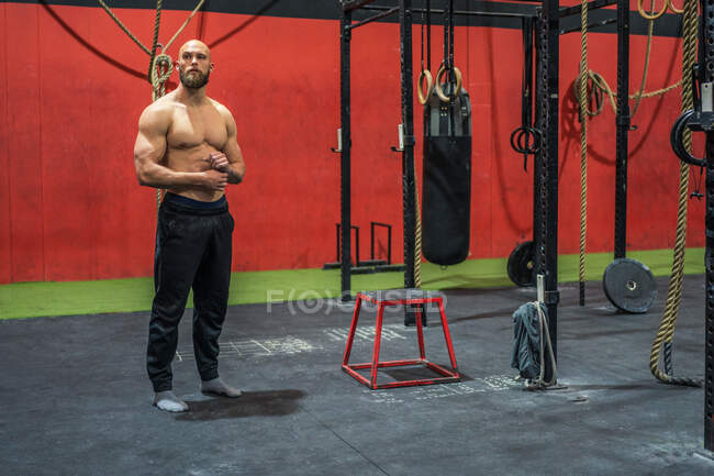 Full body powerful bearded male looking away and thinking while standing near equipment during training in modern gym — Stock Photo
