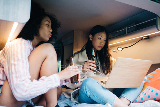 Jovens amigas multirraciais bebendo cerveja e usando laptop juntos enquanto preparam conteúdo para blog de viagens dentro van campista durante aventura de verão na natureza — Fotografia de Stock