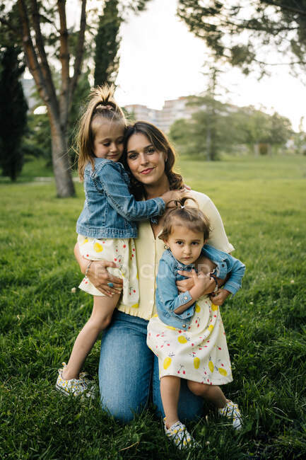 Happy young mother with cute little daughters in similar clothes enjoying sunny summer day together while sitting on blanket on grassy lawn in park — Stock Photo