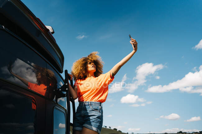 Dal basso donna nera con i capelli ricci in piedi sulla scala fuori furgone e prendendo selfie contro cielo blu nuvoloso durante il viaggio in auto — Foto stock
