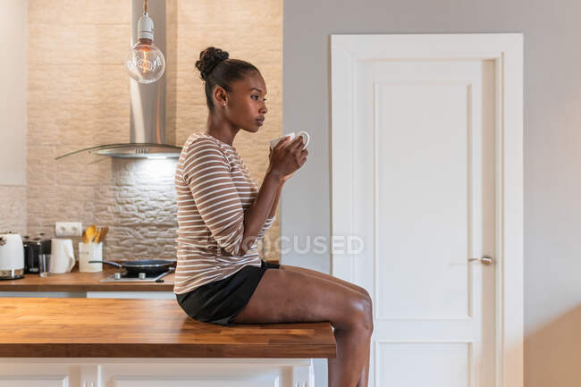 Side view of young dreamy African American female with mug of hot drink sitting on table and looking away indoors — Stock Photo