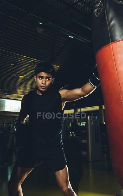 Молодой ориентированный азиат тренирует бокс выполняя удары, тренируясь с тяжелой боксерской грушей в современном тренажерном зале — стоковое фото
