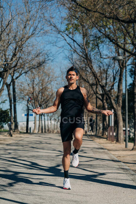 Male athlete in sportswear jumping with skipping rope and looking away on walkway during cardio training in park — Stock Photo