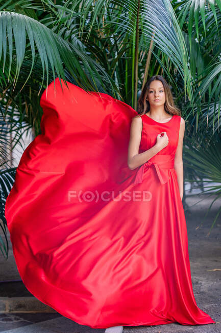Corps complet de mannequin adolescente romantique aux cheveux longs portant une robe maxi rouge vif debout près de plantes tropicales vertes et regardant la caméra — Photo de stock