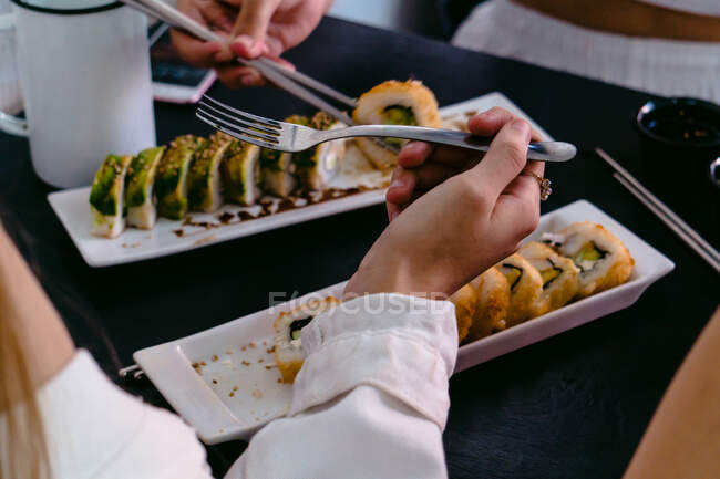 High angle of crop anonymous friends with reusable chopsticks and fork eating delicious sushi rolls with soy sauce and jalapeno pepper — Stock Photo