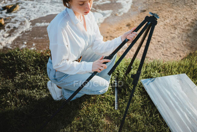 From above young stylish female painter in casual clothes and beret setting up easel on grassy terrain above sandy beach of wavy ocean on sunny day — Stock Photo