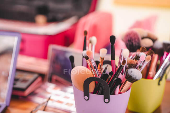 Focus morbido della tazza con pennelli trucco assortiti posizionati sulla tavola con cosmetici in salone — Foto stock