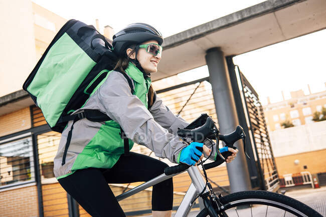 Low angle of female courier with thermal bag smiling and riding bike on street while making delivery on sunny day in city — Stock Photo