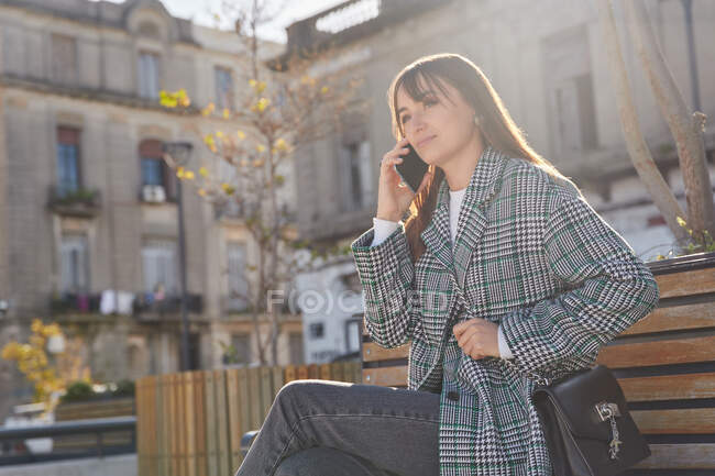 Moderna hembra milenaria con elegante atuendo de primavera sentada en el banco y respondiendo a una llamada telefónica mientras descansa en la calle urbana mirando hacia otro lado - foto de stock