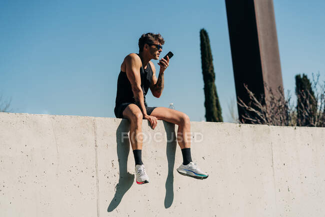 Fit male athlete in sports clothes sending voice message on cellphone while resting on fence after workout — Stock Photo