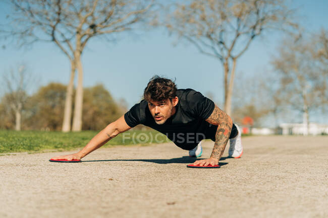 Nível do solo do atleta masculino atencioso na prancha posar com discos de deslizamento de treinamento e olhando para a frente no pavimento na cidade — Fotografia de Stock