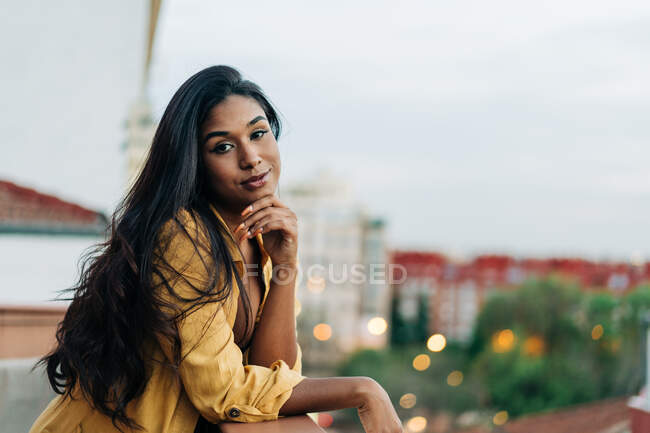 Giovane donna ispanica felice in abiti casual appoggiata sulla ringhiera e guardando la fotocamera mentre si rilassa sul balcone in serata in città — Foto stock