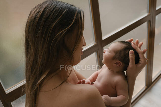 Side view of tender mother with closed eyes standing with cute naked infant near window at home — Stock Photo