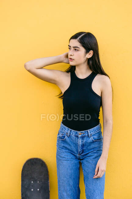 Young female skater with skateboard standing looking away on walkway with colorful yellow wall on the background in daytime — Stock Photo