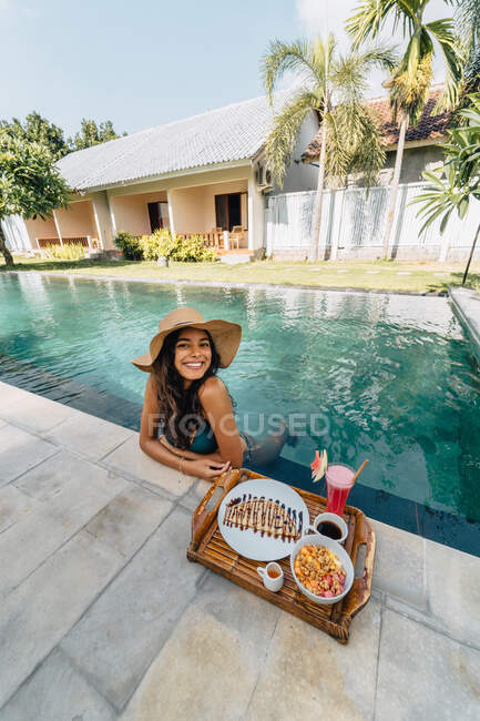 Alegre turista femenina apoyada en la piscina mientras mira a la cámara contra la bandeja con delicioso desayuno a la luz del sol - foto de stock
