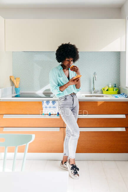 Jovem afro-americana em uso casual comer maçã e navegar na internet no telefone celular, enquanto em pé na cozinha em casa — Fotografia de Stock