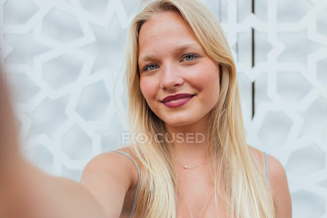 Mulher alegre com cabelo loiro olhando para a câmera ao tomar auto-tiro no telefone celular na rua da cidade — Fotografia de Stock