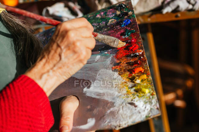 Desde arriba de la cosecha irreconocible pintor senior mezcla de colores en la paleta de pintura en el taller de arte - foto de stock