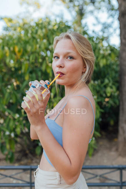 Vista lateral de encantadora hembra bebiendo limonada refrescante fría con paja mientras se enfría en el jardín de verano y mirando a la cámara - foto de stock