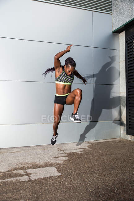 Femme afro-américaine musclée en vêtements de sport regardant la caméra et sautant tout en travaillant sur la rue de la ville près du mur de bâtiment moderne — Photo de stock