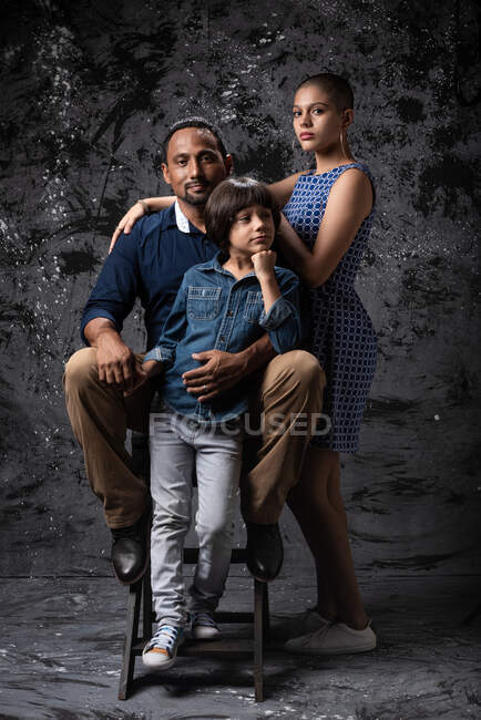 Stilvolles multirassisches Paar mit Teenager-Sohn, das im Studio auf dunklen Hintergrund in die Kamera blickt — Stockfoto