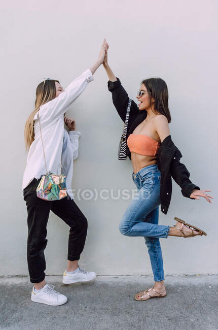 Full body side view of cheerful girlfriends wearing stylish outfits raising hands and doing high five — Stock Photo