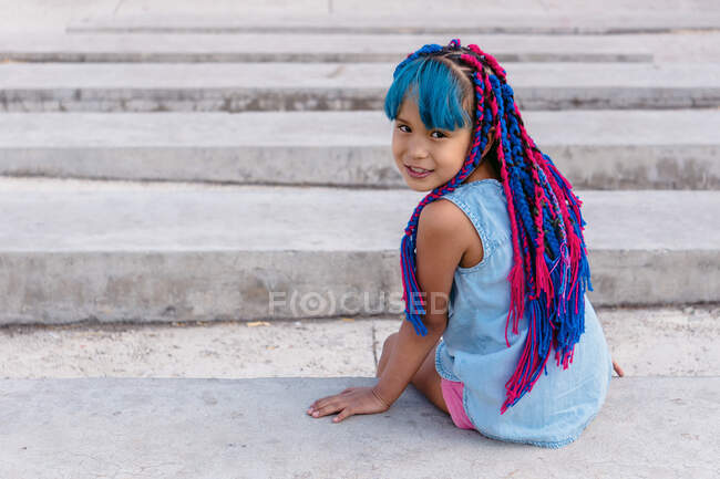 Joyeux enfant ethnique mignon avec des tresses colorées assis sur un escalier en béton tout en regardant la caméra en plein jour — Photo de stock