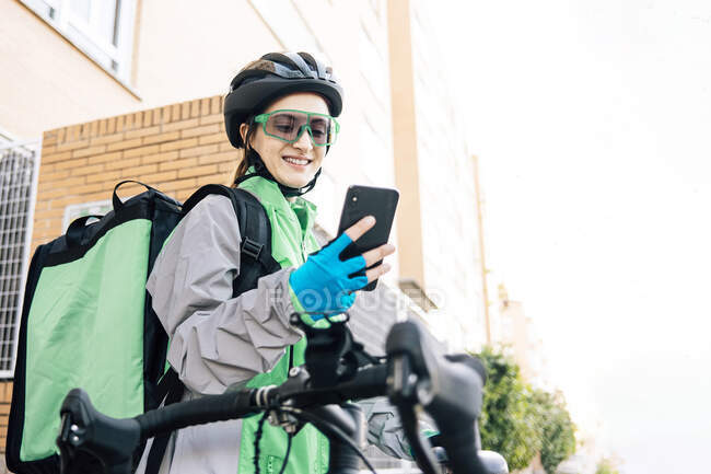 Jovem mulher alegre com saco térmico que navega smartphone com mapa GPS enquanto está sentado na bicicleta para entregar comida na rua da cidade — Fotografia de Stock