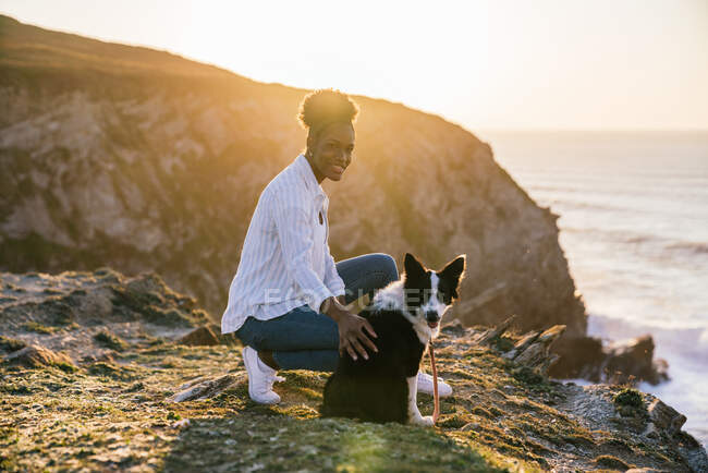 Бічний вид на молоду афро-американську жінку власницю з прикордонником Коллі Пес проводить час разом на пляжі біля махання морем на заході сонця, роздумуючи над видом — стокове фото