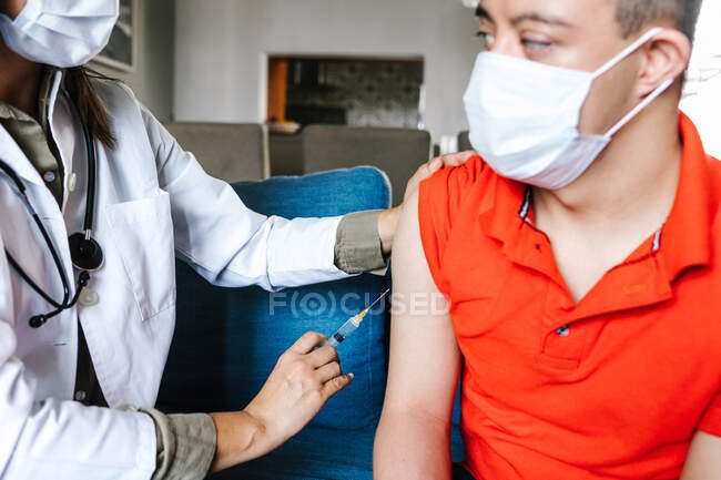 Médico fêmea da colheita com seringa que faz a injeção da vacina para o menino adolescente latino com síndrome de Down em casa durante o coronavírus — Fotografia de Stock