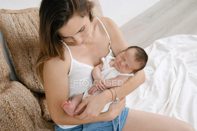 De cima delicada mãe sentada com bebê adorável em cama macia no quarto acolhedor em casa — Fotografia de Stock