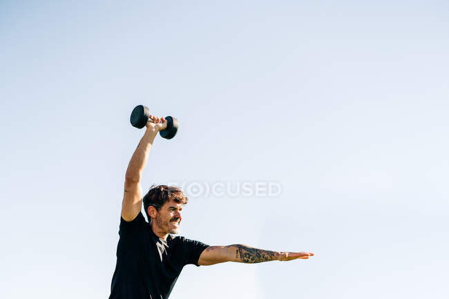 Concentrated male athlete in sportswear working out with dumbbell while looking forward under light sky — Stock Photo