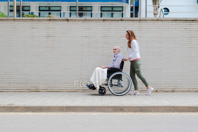 Side view of adult daughter pushing wheelchair with elderly father while walking together in city — Stock Photo