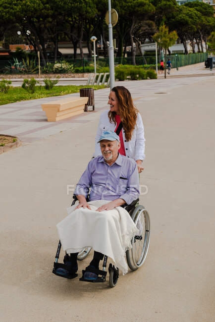 Mujer adulta sonriente y hombre mayor en silla de ruedas relajándose juntos en la ciudad en un día soleado - foto de stock