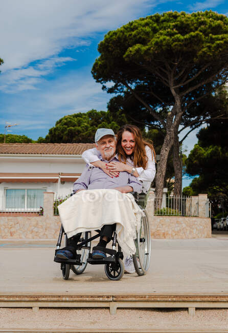 Mujer adulta sonriente y hombre mayor en silla de ruedas relajándose juntos en la ciudad en un día soleado - foto de stock